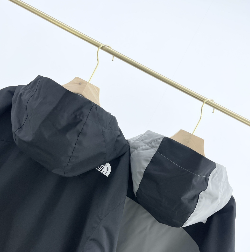 Two hooded outdoor jackets in black and grey hanging on wooden hangers against a white background.